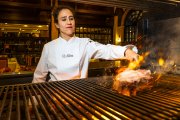 Vanesa Martín maneja las llamas de su flamante parrilla en El Alfoz para cocinar una chuleta. Tras ella, el salón en el que los comensales pueden apreciar su trabajo.