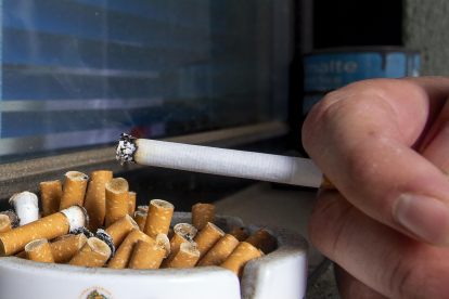 Detalle de un cenicero lleno de colillas mientras una persona sostiene un cigarro. SANTI OTERO