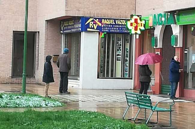 Compradores hacen cola frente a una farmacia esta mañana bajo la lluvia esperando a ser atendidos, dejando más de un metro de distancia. S. L. C.