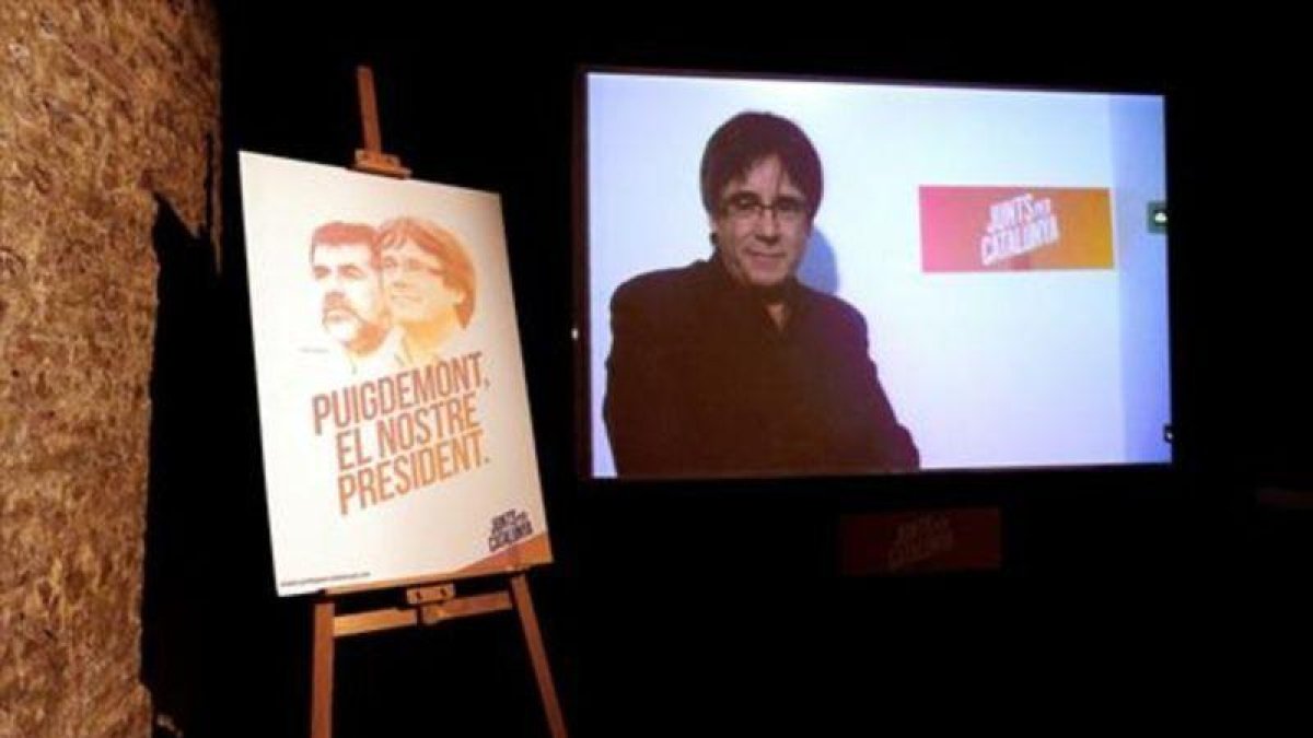 Videoconferencia de Puigdemont, junto a un cartel de Junts per Catalunya.-EFE / ALBERTO ESTÉVEZ