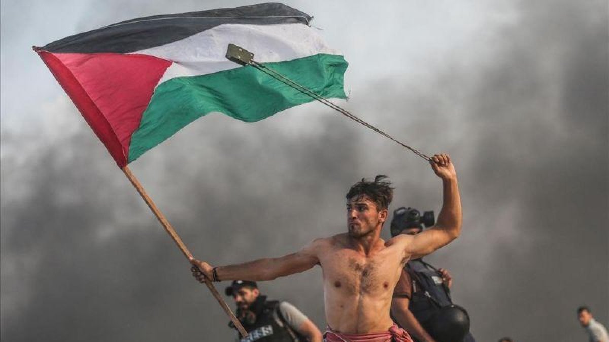 Un joven palestino tira piedras con una honda mientras con la otra mano lleva la bandera palestina.-MUSTAFA HASSONA / GETTY IMAGES