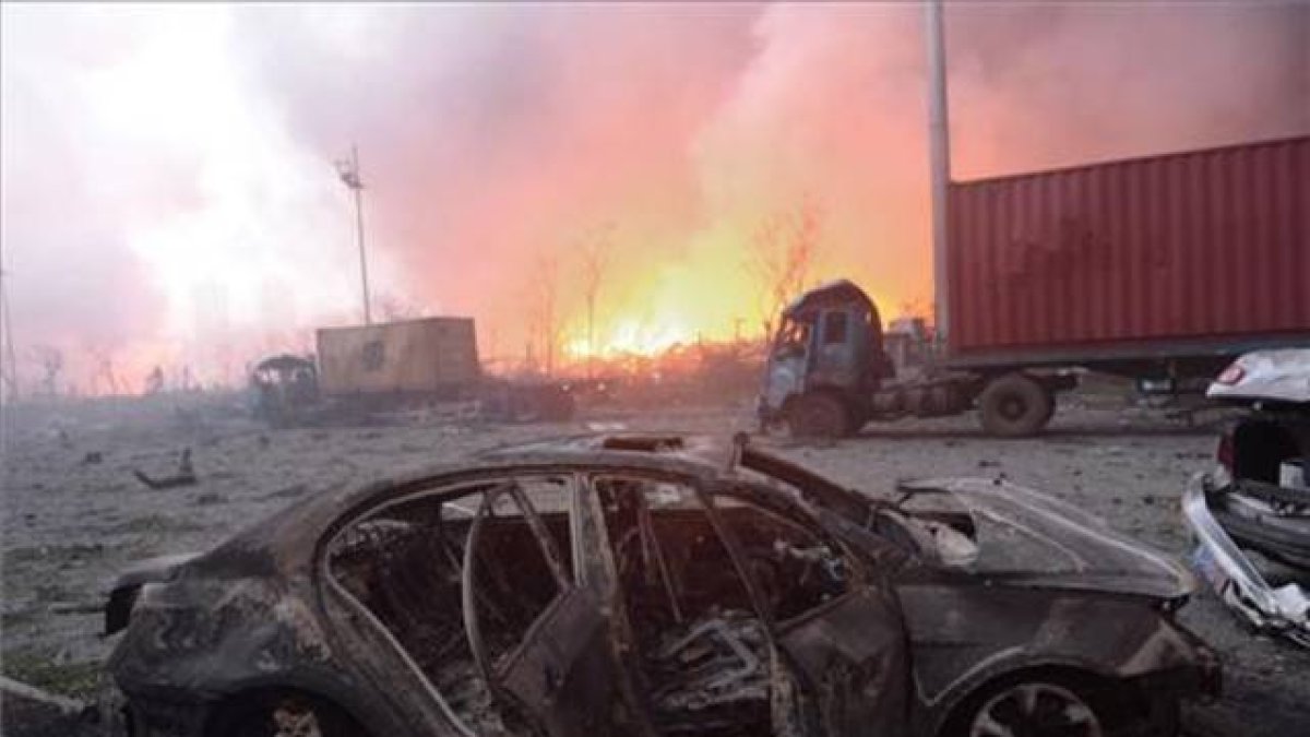 Vista de un coche calcinado en la seria de explosiones de unos almacenes de Tianjin, en China. En segundo plano, el fuego sigue ardiendo.-AFP / TIANJIN