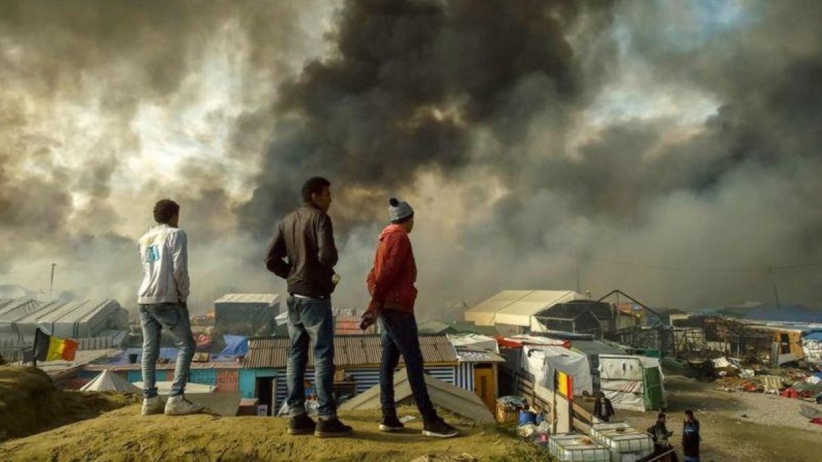 Inmigrantes observan el campamento evacuado de Calais con parte de las barracas ardiendo.-AFP / PHILIPPE HUGUEN