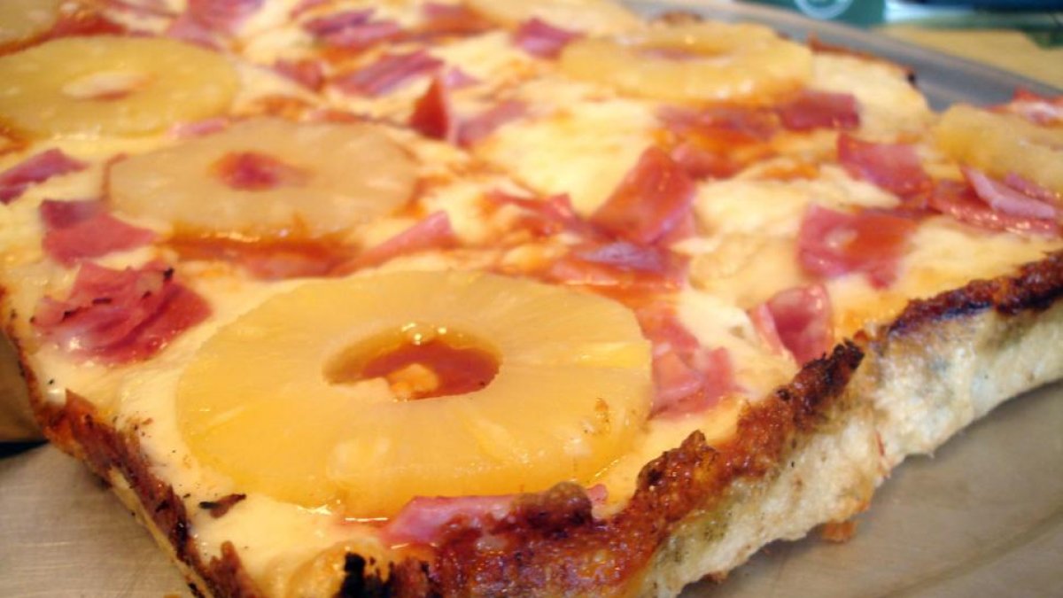 Una porción de pizza hawaiana, con rodajas de piña enteras.-
