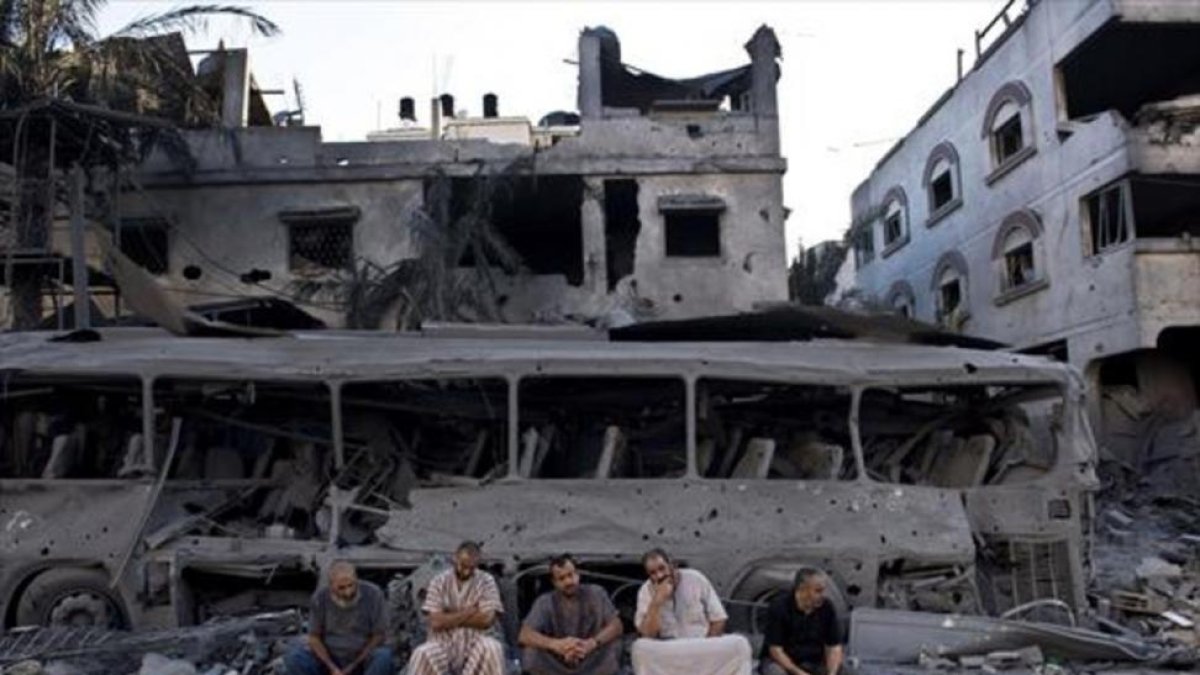 Cinco palestinos de Gaza sentados frente a un autobús y un edificio destruidos, en el 2014, por ataques aéreos israelís.-AFP / ROBERTO SCHMIDT