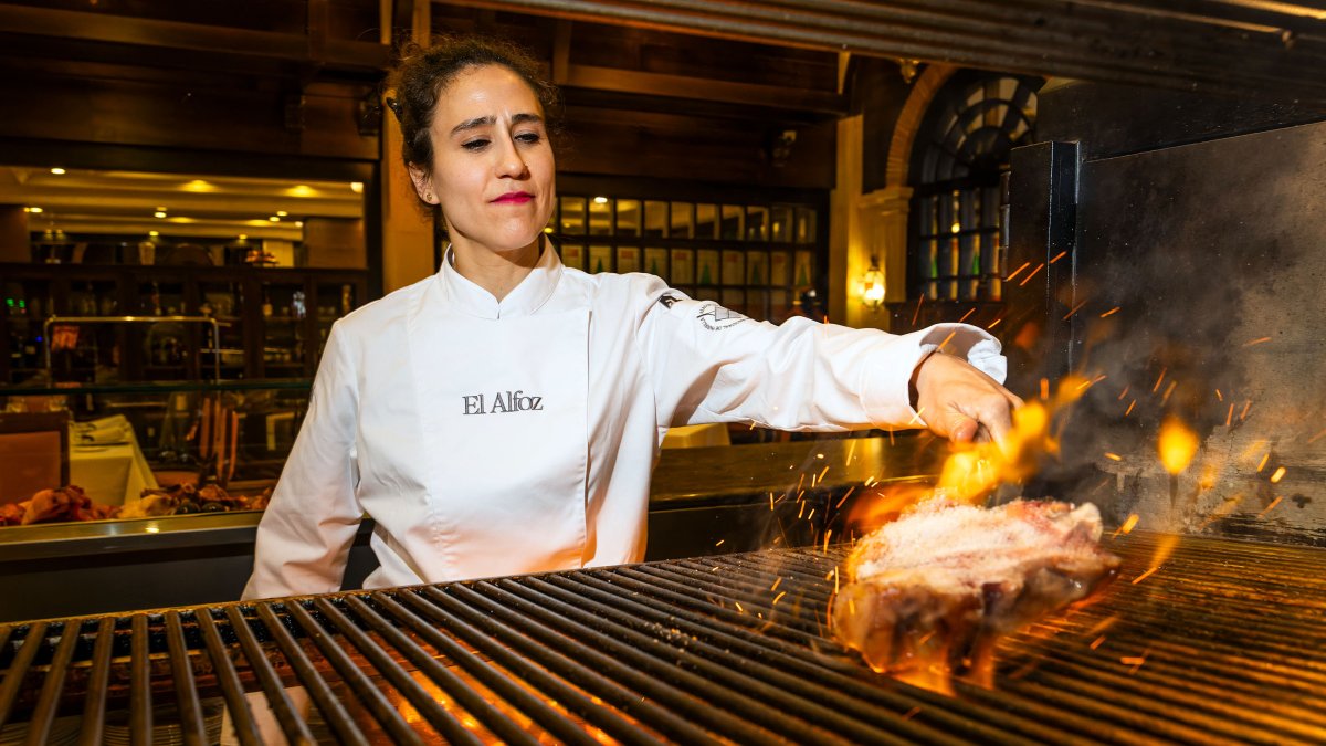 Vanesa Martín maneja las llamas de su flamante parrilla en El Alfoz para cocinar una chuleta. Tras ella, el salón en el que los comensales pueden apreciar su trabajo.